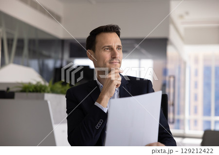 Focused man holding documents, gazes thoughtfully into distance 129212422