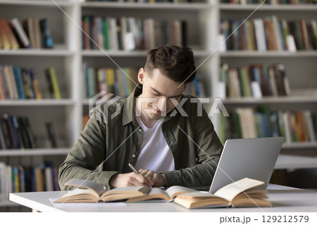Smiling male student write academic work using books and laptop Smiling male student write academic work using books and laptop 129212579