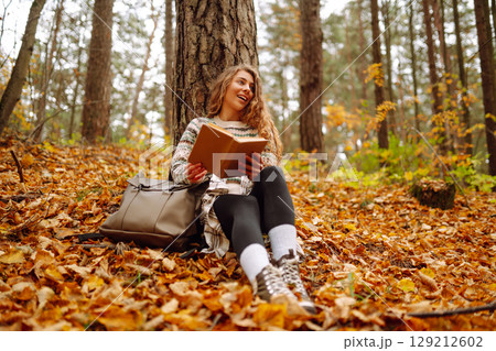 Curly-haired woman in a stylish sweater with a book sits under a maple tree. Concept of autumn. 129212602