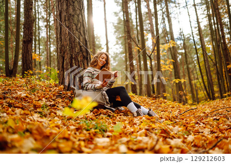 Curly-haired woman in a stylish sweater with a book sits under a maple tree. Concept of autumn. 129212603