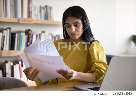 Businesswoman reviewing printed documents while sit at desk with laptop 129212842