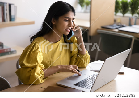 Serious Indian woman speak on phone while working on laptop 129212885