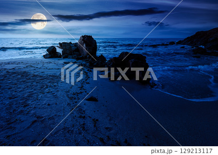 landscape by the sea at night in summer. beautiful beach with rocks washed by calm waves in full moon light. view to horizon under dark sky with clouds. scenic travel destination for leisure and relax 129213117
