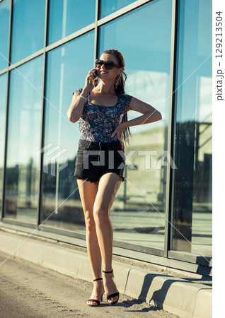 A girl wearing shorts, a T-shirt and a ponytail is on the phone in front of a glass facade 129213504