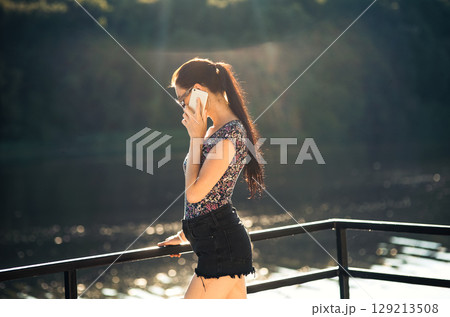 The young girl stands by the lake, holding her phone and smiling at a joke 129213508