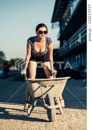 A pretty girl in summer clothes stands by scaffolding, resting her hand on the handle of a wheelbarrow 129213557