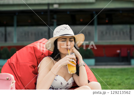 A stylish girl in a hat is sitting on a red beanbag chair and slowly drinking juice 129213562