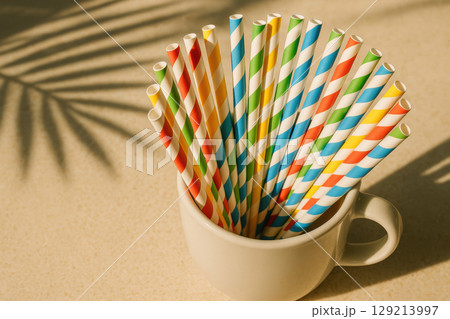 Colorful striped paper drinking straws arranged in white ceramic mug with sunlight and palm leaf shadow on beige background, AI Generative 129213997