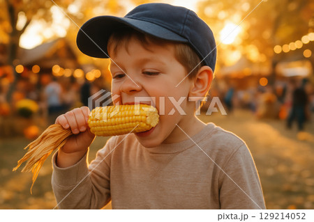 Young boy enjoys eating fresh corn on the cob outdoors during an autumn festival with warm golden sunlight in the background, AI Generative 129214052