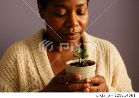 Calm woman holding small green seedling in ceramic pot, nurturing indoor plant growth with gentle hands against soft purple background, AI Generative 129214061