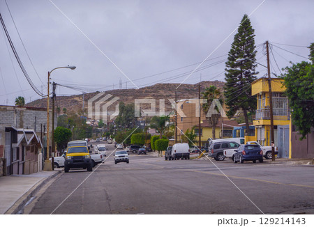 Street in Tijuana Colonia Libertad with modest houses, parked cars, power lines and hills beneath overcast sky. 129214143