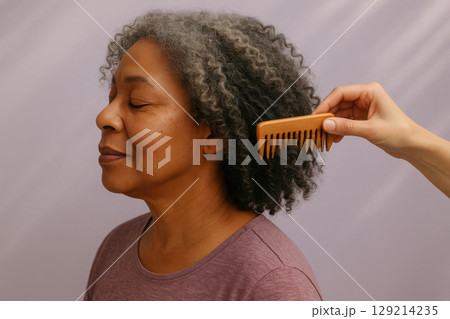 Smiling mature woman with natural curly gray hair being gently combed with a wide tooth wooden comb against neutral background, AI Generative 129214235