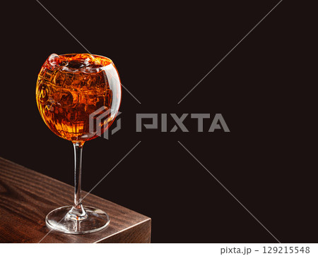 A cocktail with ice served in a large stemmed glass on a dark wooden bar with a black background. copy space for text 129215548