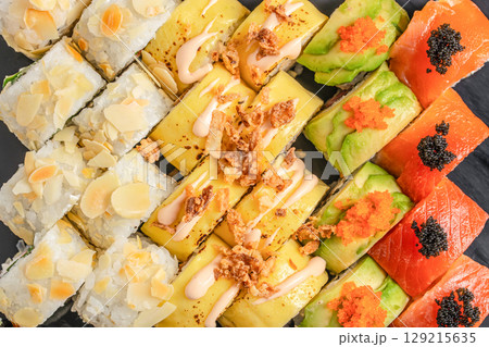 An assorted platter of vibrant sushi rolls topped with almonds, mango, avocado, and salmon with colorful roe, beautifully arranged on a dark slate background An assorted platter of vibrant sushi rolls topped with almonds, mango, avocado, and salmon with colorful roe, beautifully arranged on a dark slate background 129215635