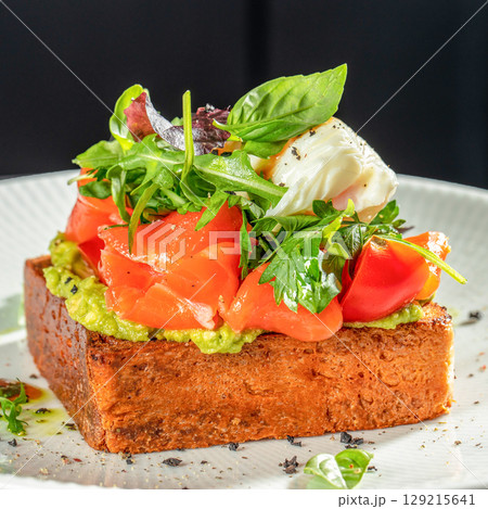 Gourmet toast topped with avocado spread, smoked salmon, cherry tomatoes, mixed greens, and a poached egg on a white plate Gourmet toast topped with avocado spread, smoked salmon, cherry tomatoes, mixed greens, and a poached egg on a white plate 129215641