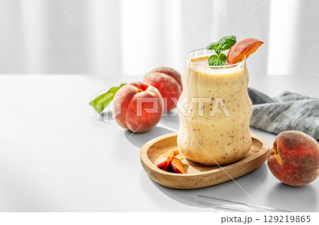 Smoothie with peach and banana in a glass on a wooden board on a white table near window 129219865