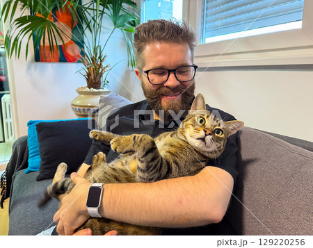 Happy man holding his cat on a couch at home. Companionship, relaxation, and emotional comfort through the bond between human and pet. Happy man holding his cat on a couch at home. Companionship, relaxation, and emotional comfort through the bond between human and pet. 129220256