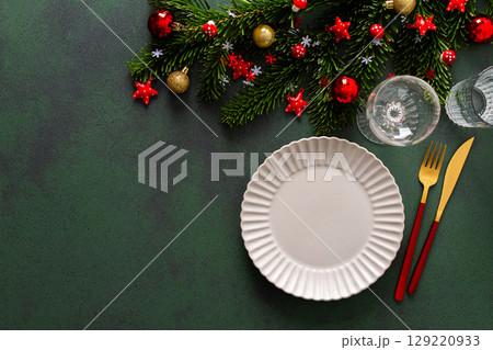 Elegant Christmas table setting with an empty plate and cutlery on a deep green surface. Red Christmas decorations and spruse branches add a warm, holiday touch Elegant Christmas table setting with an empty plate and cutlery on a deep green surface. Red Christmas decorations and spruse branches add a warm, holiday touch 129220933