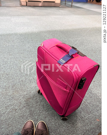Pink carry-on suitcase at airport gate with visible shoes in frame. Travel preparation, mobility, and passenger experience in transit environment Pink carry-on suitcase at airport gate with visible shoes in frame. Travel preparation, mobility, and passenger experience in transit environment 129221127