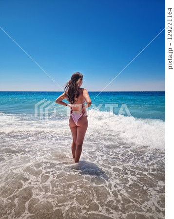 Rear view beautiful woman in bikini stands in the foamy waves of the clear blue sea, facing the wide horizon under a cloudless summer sky. Summertime vacation on a tropical beach, relaxation vibes Rear view beautiful woman in bikini stands in the foamy waves of the clear blue sea, facing the wide horizon under a cloudless summer sky. Summertime vacation on a tropical beach, relaxation vibes 129221164
