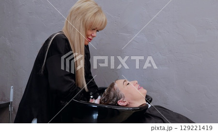 Professional hairdresser washing client's hair with shampoo and water in a black sink, providing a relaxing and pampering experience in a salon setting 129221419