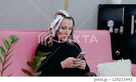 Young woman with foil in her hair uses a smartphone while waiting for her hair dye to set in at a beauty salon, relaxing on a pink sofa Young woman with foil in her hair uses a smartphone while waiting for her hair dye to set in at a beauty salon, relaxing on a pink sofa 129221443