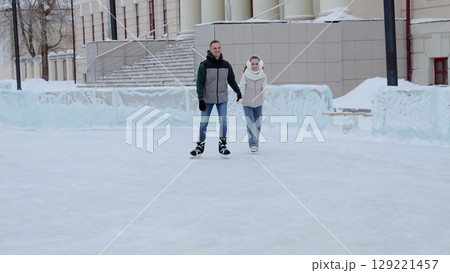 Loving couple gliding together on frozen rink, holding hands during wintertime recreational skating, sharing intimate moment against snowy urban landscape 129221457