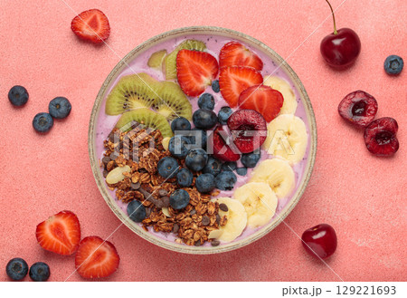 Delicious acai bowl with fresh fruits and granola in white ceramic bowl on pink table top view, healthy breakfast or snack 129221693