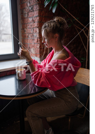 A stylish woman enjoying her coffee while scrolling on her phone in a warm cafe setting. 129221765