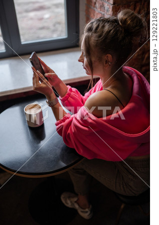A stylish woman enjoying her coffee while scrolling on her phone in a warm cafe setting. A stylish woman enjoying her coffee while scrolling on her phone in a warm cafe setting. 129221803