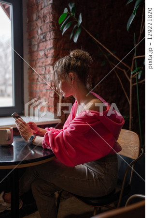 A stylish woman enjoying her coffee while scrolling on her phone in a warm cafe setting. 129221820