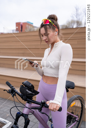 A woman in stylish activewear checks her phone, showcasing the vibrant outdoor fitness culture by her bicycle. A woman in stylish activewear checks her phone, showcasing the vibrant outdoor fitness culture by her bicycle. 129221848