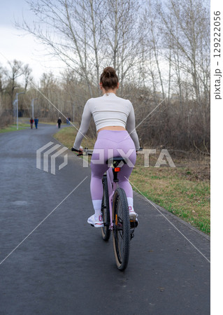 Young woman riding a bicycle in the park on the background of trees. Trendy active girl spends time on the nature. Side view. 129222056