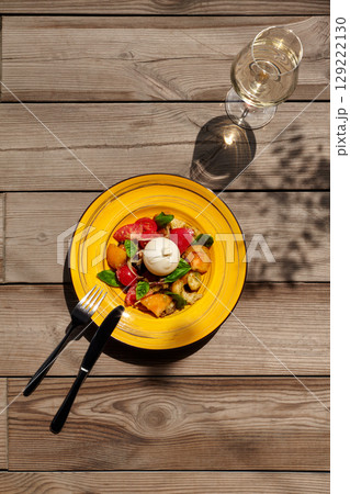 Panzanella salad with burrata cheese, tomatoes, and basil enjoying sunlight on wooden table 129222130