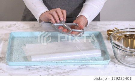 A flat beater attachment for a kitchen mixer is held above a parchment-lined tray, preparing for a baking session. The background includes measuring cups and a glass mixing bowl. 129225401