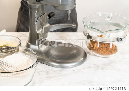 Eye-level view of a stand mixer with a glass bowl, blending the ingredients for Classic Gingerbread Cookie Dough. Visible layers include the dark molasses mixture and brown sugar, ready to be combined 129225418