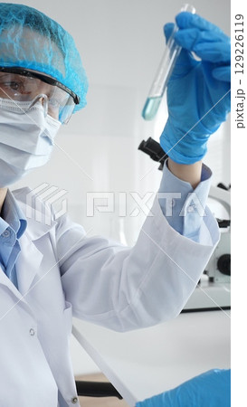Close up of a scientist in ppe, holding a test tube with blue liquid while conducting research in a laboratory with a microscope. Medicine, health care and science concept Close up of a scientist in ppe, holding a test tube with blue liquid while conducting research in a laboratory with a microscope. Medicine, health care and science concept 129226119