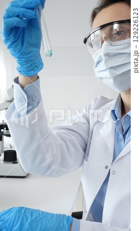Female scientific researcher holding test tube, analyzing blue liquid while wearing protective gear during medical experiment in sterile laboratory setting. Medicine and science concept 129226123
