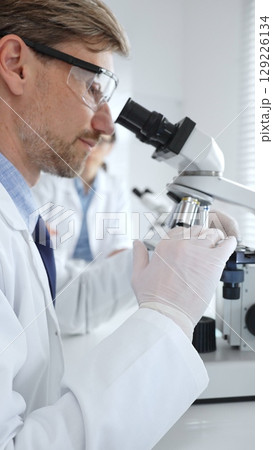 Researcher examining sample through microscope, wearing safety eyewear in sterile laboratory setting, surrounded by scientific glassware. Medicine, healthcare and science concept 129226134