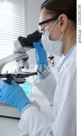 Female scientist wearing mask and blue gloves using microscope doing scientific research in modern laboratory with colleague working in background. Medicine, healthcare and science concept Female scientist wearing mask and blue gloves using microscope doing scientific research in modern laboratory with colleague working in background. Medicine, healthcare and science concept 129226136