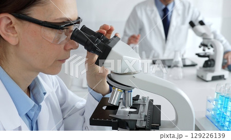 Female scientist examining specimen under microscope, conducting research in sterile laboratory with colleague working near test tube rack. Medicine, healthcare and science concept 129226139