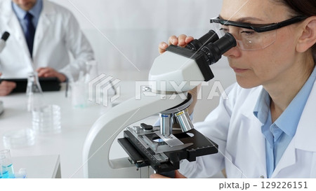 Female scientist examining sample under microscope, conducting research in laboratory with colleague working near test tube rack. Concept of medicine, healthcare and science Female scientist examining sample under microscope, conducting research in laboratory with colleague working near test tube rack. Concept of medicine, healthcare and science 129226151