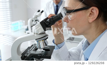 Female scientist examining sample under microscope, conducting research in laboratory with colleague working near test tube rack. Concept of medicine, healthcare and science Female scientist examining sample under microscope, conducting research in laboratory with colleague working near test tube rack. Concept of medicine, healthcare and science 129226208