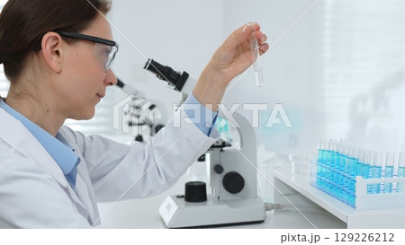 Female researcher examining clear liquid in test tube, sitting near microscopes and lab equipment within sterile scientific workspace. Medicine, healthcare and science concept Female researcher examining clear liquid in test tube, sitting near microscopes and lab equipment within sterile scientific workspace. Medicine, healthcare and science concept 129226212