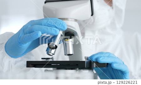 Close up of scientific researcher or virologist wearing protective gear is adjusting microscope, analyzing lab samples with precision in sterile research environment. Medicine and science concept Close up of scientific researcher or virologist wearing protective gear is adjusting microscope, analyzing lab samples with precision in sterile research environment. Medicine and science concept 129226221