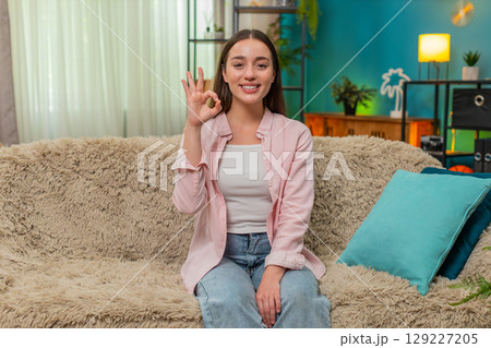 Young woman making confident okay hand gesture smiling to camera showing approval and trust on sofa 129227205