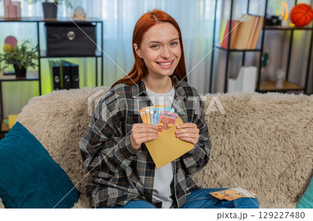 Woman sitting on sofa counting cash euro putting part into envelope looking happy and satisfied 129227480