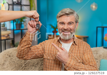 Mature man on sofa receiving keys to new apartment from realtor, smiling after lottery win at home 129227519