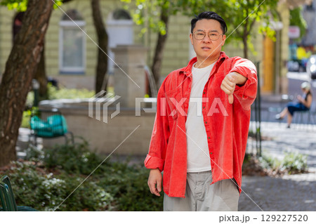 Upset Asian man showing thumbs down sign, disapproval, dissatisfied bad work, mistake in city 129227520