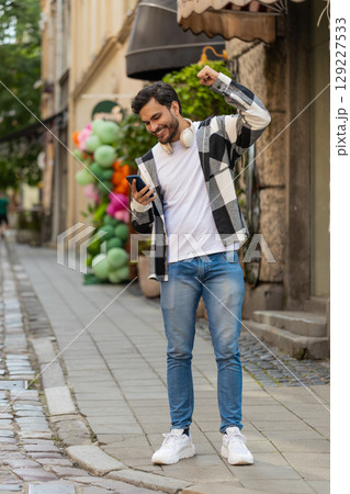 Hispanic young man using smartphone celebrating win good message news clenching fist on city street 129227533